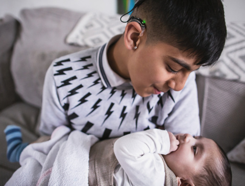 a boy wearing a hearing aid, holding his baby sister and looking at each other