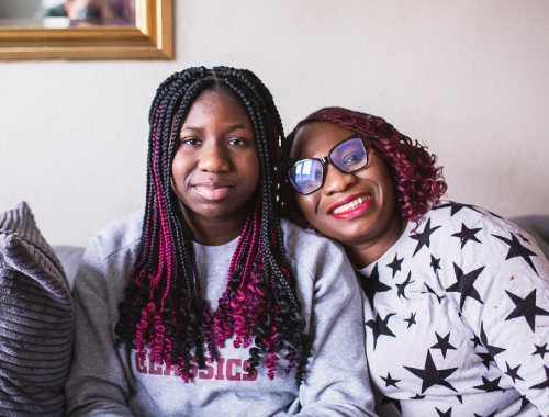 A teenage girl with long hair, and her mum seated next to here on a sofa smiling