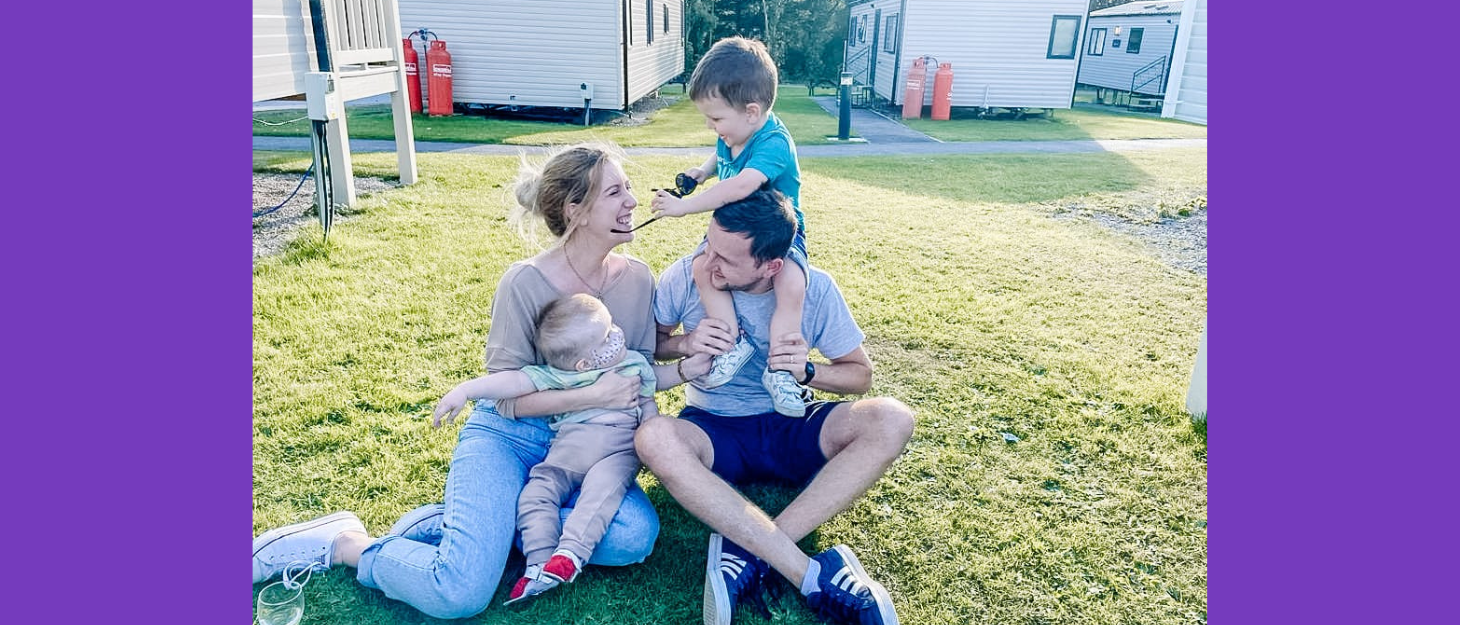 Family sit on grass outside caravan park. Mum holds one twin on her lap and dad has the other twin on his shoulders.