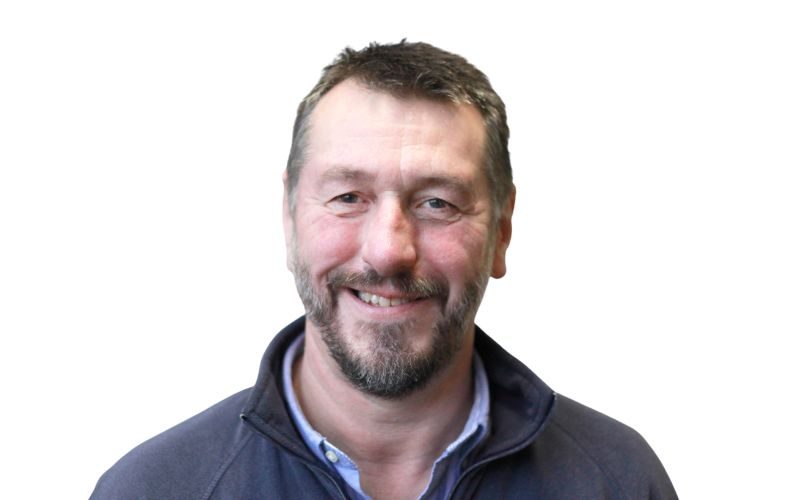 A man with a shirt and navy pull over, dark hair and short beard