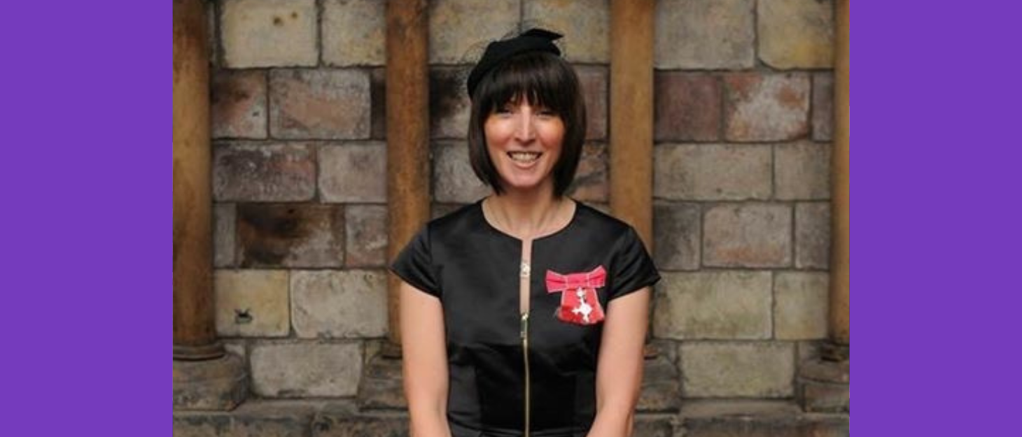 A woman with short dark hair wearing her MBE in a black dress and hat, she's smiling