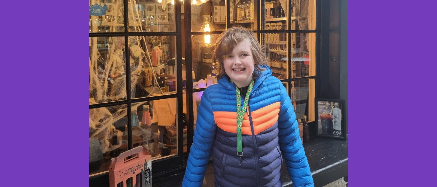 Boy wearing rain coat and sunflower lanyard stands outside magic shop smiling at camera