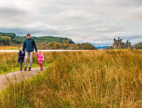 A dad walking with two small children across a field in Scotland with a castle in the background