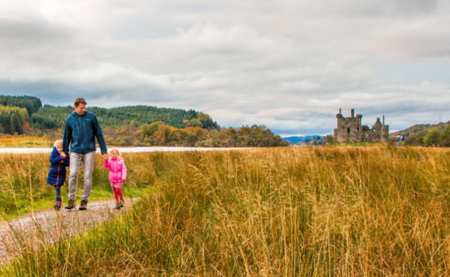 A dad walking with two small children across a field in Scotland with a castle in the background