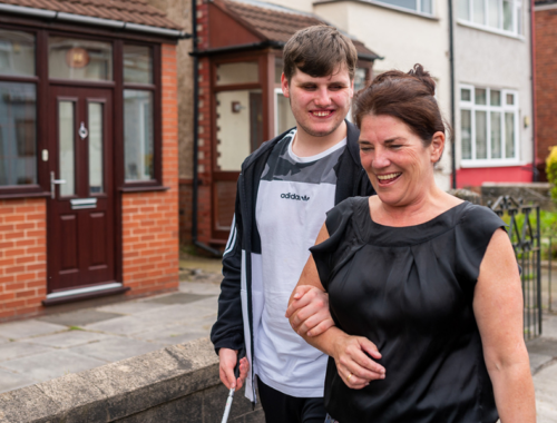 A mum and her son walk in the street, with arms linked. She has a sleeveless top and is laughing. He is smiling at her and has a walking stick