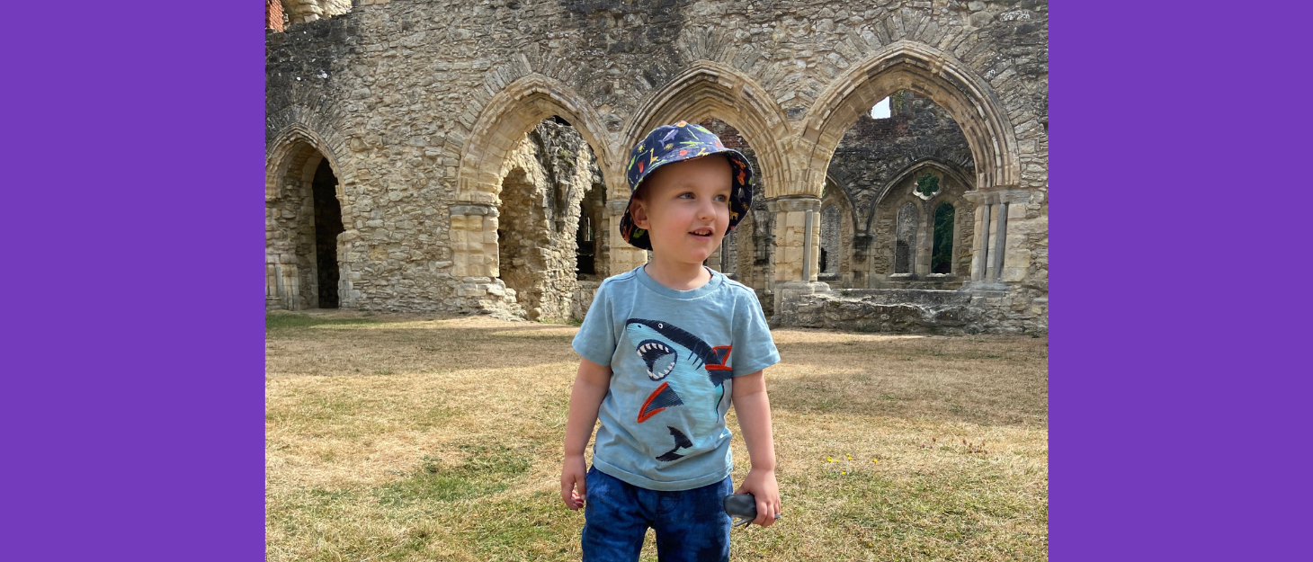 Young boy wearing teeshirt and hat stands in front of old ruin on sunny day