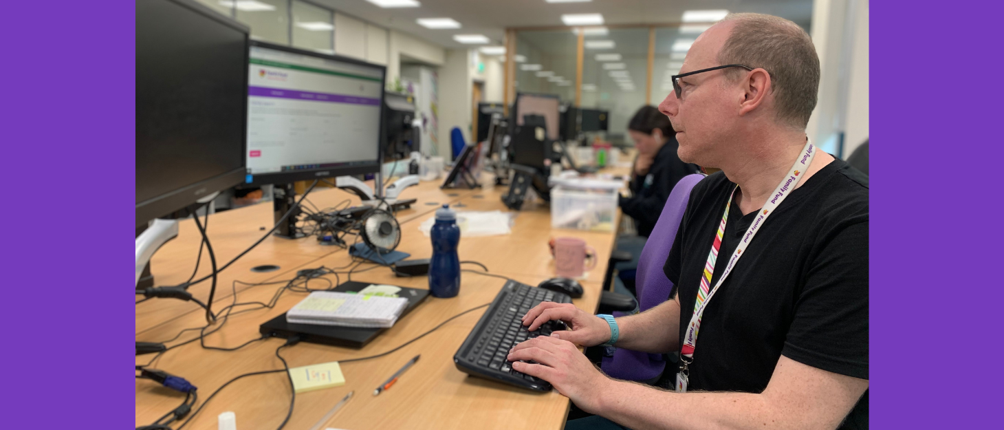 A man, wearing a black tee-shirt and a family fund lanyard is sitting in an office, and working on a computer