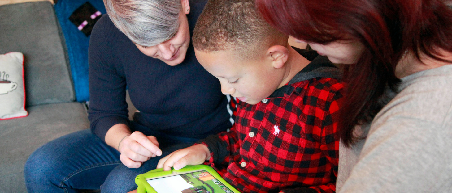 A boy sits with his mum and sister. He is using an iPad.