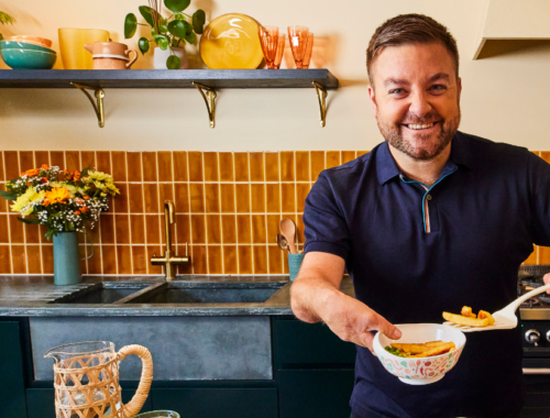 Comedian Alex Brooker stood in a kitchen holding a limited edition scoop bowl in one hand an a spatula with chips on in the other