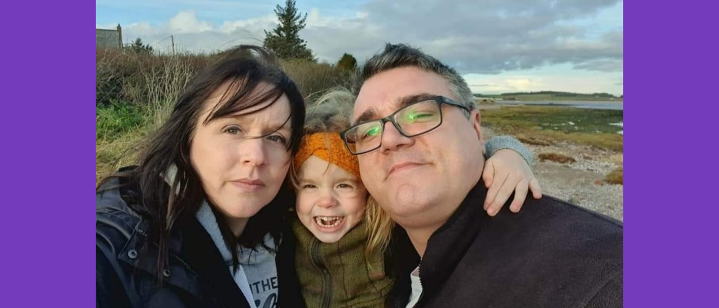 A little girl cuddles between her mum and dad, and they all pose for a photo out on a walk